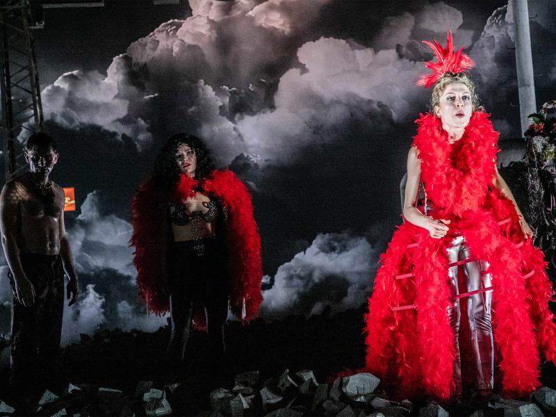 Alberta von Poelnitz im Vordergrund, sie trägt roten Feder-Kopfschmuck und eine rote Federboa um die Schultern, die in einem mit roten Federn behängten Reifrock mündet. Ihr Mund ist geöffnet, sie schaut nach vorn. Hinter ihr im Dunkeln stehen Paul Behren mit nacktem Oberkörper und Linn Reusse, ebenfalls mit roter Federboa, einem mit Strass besetzten knappen BH und langer Hose. Der Boden ist mit Kohle bedeckt.