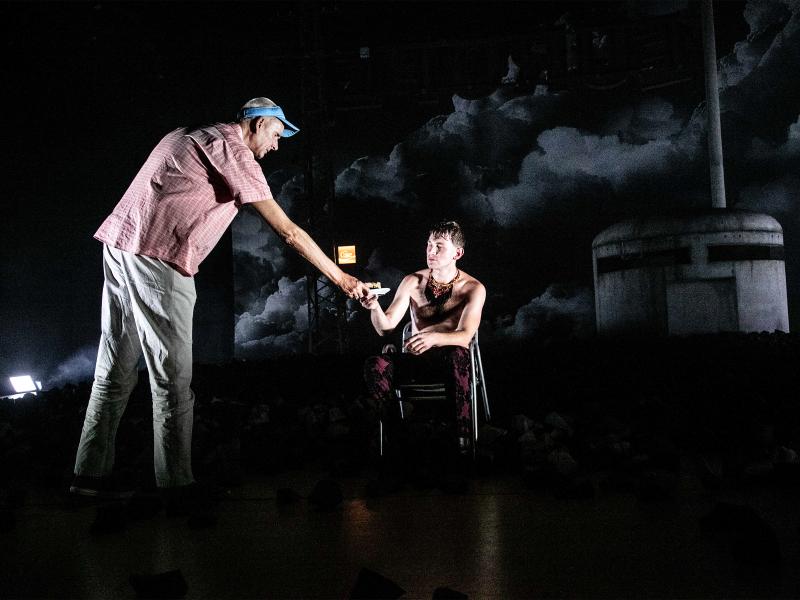 Olaf Rausch mit heller Hose, rot kariertem Hemd und einem hellblauen Sonnenvisier auf dem Kopf. Er reicht Paul Behren einen Teller, der mit nacktem Oberkörper und roter Hose auf einem Metallstuhl sitzt. Im Hintergrund ein graues rundes Gebäude aus schmutzigem Beton mit großen horizontalen Schlitzen.