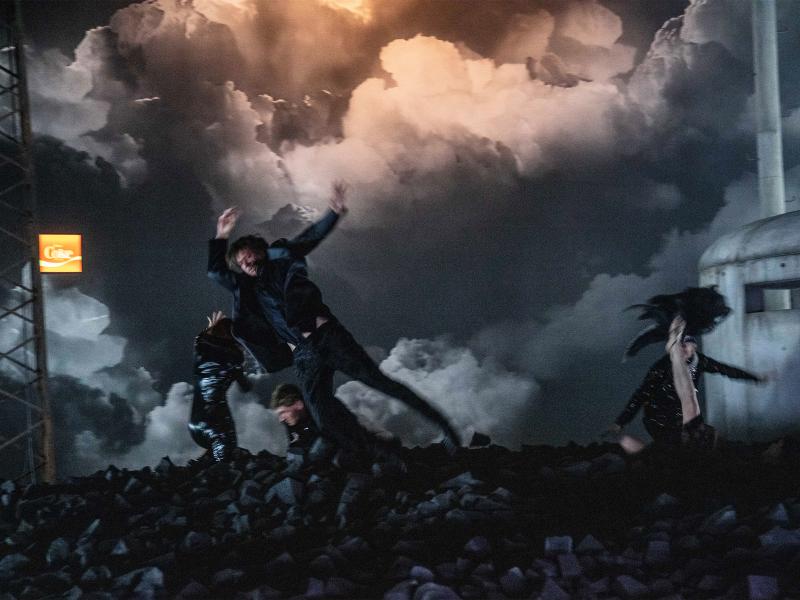 Vier schwarz gekleidete Personen springen quer durch aufgehäufte Kohlestücken am Boden.