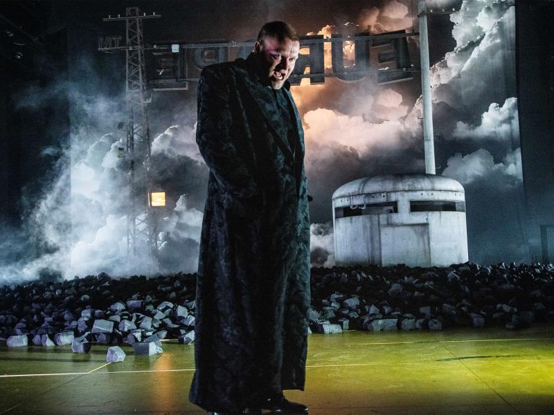 Eine Person mit bodenlangem schwarz-gemustertem Mantel, sein Mund ist geöffnet, er schaut nach vorn. Im Hintergrund mit großen Kohlestücken bedeckter Boden, ein graues rundes Gebäude aus schmutzigem Beton mit großen horizontalen Schlitzen und ein Strommast, an dem die Buchstaben EUROPE rückwärts umgedreht hängen. Dahinter ein bühnenfüllender Vorhang mit großen grauen Wolken.