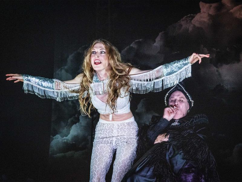 Alberta von Poelnitz mit weiß gemusterter Hose und weißem Top. Sie hat die Arme ausgebreitet, an denen silbrige Fransen hängen. Sie trägt ihr langes Haar offen, ihre Augen sind aufgerissen, ihr Mund geöffnet. Neben ihr Josef Ostendorf sitzend und schwarz mit glänzendem Stoff und schwarzen Fransen sowie einem schwarzen Hut bekleidet. Er zieht an einer Zigarette und schaut nach vorn.