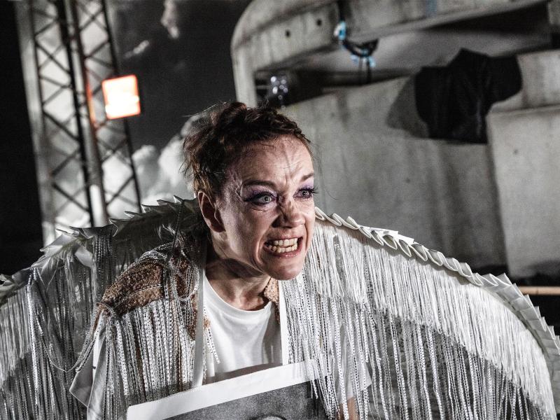 Nahaufnahme von Angelika Richter. Sie trägt ein weißes T-Shirt und eine Konstruktion mit weißen Fransen und überlappendem Stoff behängt auf den Schultern. Ihre Augen sind weit aufgerissen, ihre Zähne gefletscht. Im Hintergrund ein graues rundes Gebäude aus schmutzigem Beton mit großen horizontalen Schlitzen.