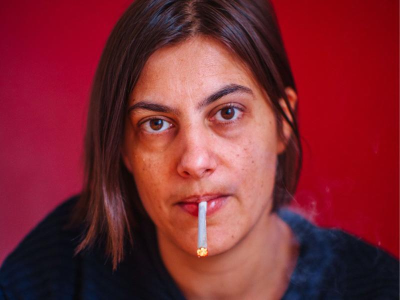 Jenny Kellner sitzt vor einer roten Wand. Sie trägt einen dunkelblauen Pullover, braune, schulterlange Haare und hat eine brennende Zigarette zwischen den Lippen. Sie schaut in die Kamera.
