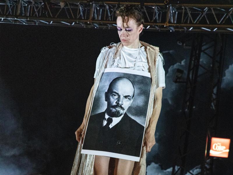 Angelika Richter im weißen T-Shirt und einem Umhang aus weißen Fransen. Sie trägt ein großes Portrait von Lenin umgehängt. Über ihr im Hintergrund eine Stahl-Traverse und ein Coke-Leuchtschild.