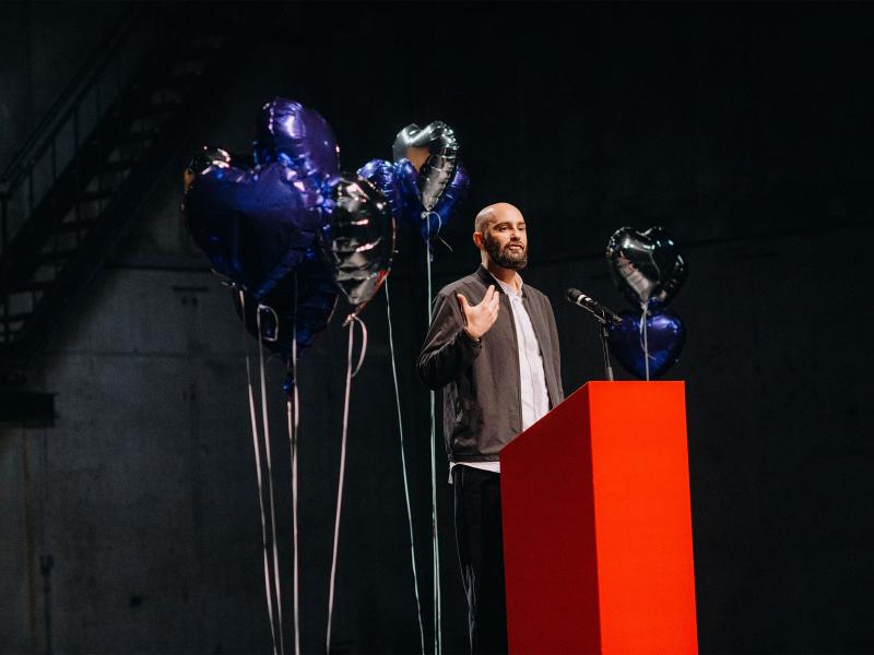 Philipp Staab steht an einem roten Podium, auf dem ein Mikrofon platziert ist, er gestikuliert mit der rechten Hand. Um ihn herum ist eine Ansammlung von silbernen und blauen herzförmigen Heliumluftballons. 