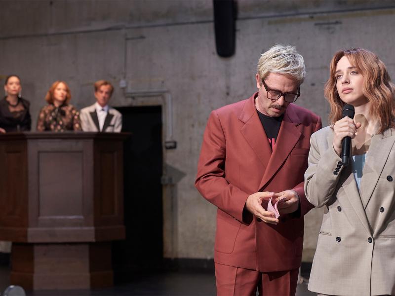 Samuel Weiss und Fee Aviv Dubois nebeneinander in einem roten und beigefarbenen Anzug im Vordergrund. Fee Aviv Dubois hält ein Mikrofon in der Hand und schaut in die Ferne, Samuel Weiss mit blonder Kurzhaarperücke und dicker Brille blättert in etwas. Im Hintergrund Sasha Rau, Josefine Israel und Magne Håvard Brekke nebeneinander auf einer hölzernen Kanzel, sie schauen zu den beiden nach vorn.