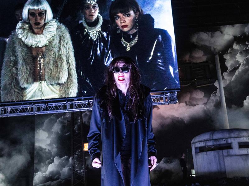 Bühnensituation: Eine Person mit langem dunklem Mantel, Sonnenbrille und langen braunen Haaren im Vordergrund. Ihr Mund ist geöffnet, die Zähne sind gefletscht. Im Hintergrund eine Leinwand mit drei weiteren Personen mit Perücken und Lack- sowie Fell-Jacke. Sie tragen ausladenden goldenen Schmuck.