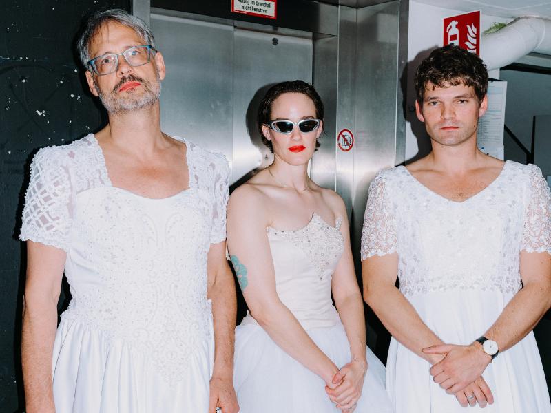 Martin Györffy, Christian Tschirner und lynn t musiol stehen in einer Reihe nebeneinander in langen weißen Kleidern. Sie posieren vor einem Fahrstuhl. 