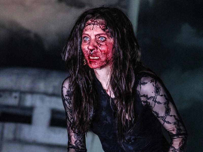 Lilith Stangenberg mit einer Perücke aus langen braunen Haaren. Ihr Gesicht ist vollständig mit Kunst blutbeschmiert, sie hat den Mund geöffnet und die Augen aufgerissen. Sie trägt ein schwarz-transparentes Oberteil.
