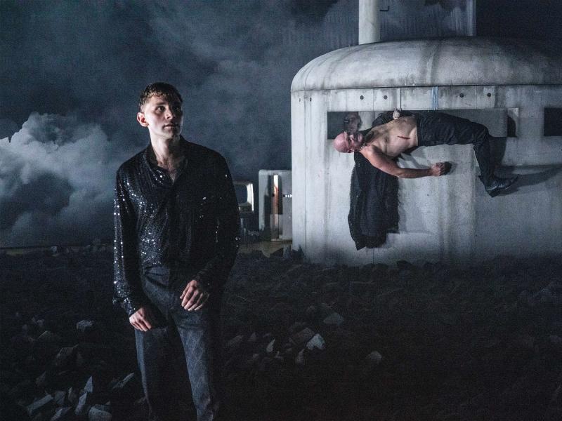 Paul Behren in einem schwarzen glitzernden Hemd und langer schwarzer Hose, er schaut in die Ferne. Im Hintergrund bedecken schwarze Kohlebrocken. Ein graues rundes Gebäude aus schmutzigem Beton mit großen horizontalen Schlitzen. In einem Schlitz liegt Matti Krause mit nacktem Oberkörper und schwarzer langer Hose, ein Bein hängt außen am Gebäude herunter.