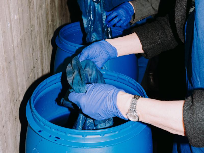 Zwei Personen tunken Tücher in drei große blaue Fässer mit blauer Farbe darin. Die Frauen tragen blaue Handschuhe und blaue Schürzen. 