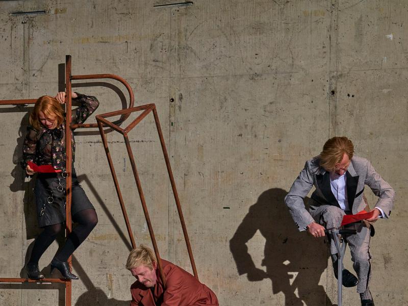 Links steht Josefine Israel auf einer abstrakte Metallkonstruktion, hält sich fest und einen roten Zettel in der Hand. Samuel Weiss, mittig, klemmt in einer dreieckigen länglichen Metallkonstruktion, es sind nur Kopf und Schultern sichtbar. Rechts Magne Håvard Brekke, er hält ebenfalls einen roten Zettel in der Hand und stützt sich mit der anderen auf eine Metallkonstruktion mit zwei Handgriffen, von der nur eine senkrechte Stange zu sehen ist.