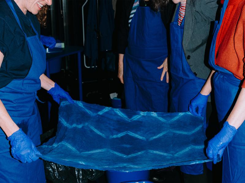 Zwei Personen in blauen Schürzen und mit blauen Handschuhen breiten gemeinsam ein blaues Tuch mit einem Muster aus. Im Hintergrund stehen weitere Personen in blauen Schürzen. 