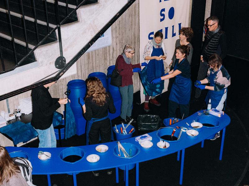Viele Menschen stehen verteilt im MalerSaal Foyer, einige halten blaue Tücher in den Händen. In der Mitte des Raumes ist ein blauer langer Tisch, auf dem sich Werkzeuge und Bastelutensilien befinden. Es sind Eimer in den Tisch eingelassen. 