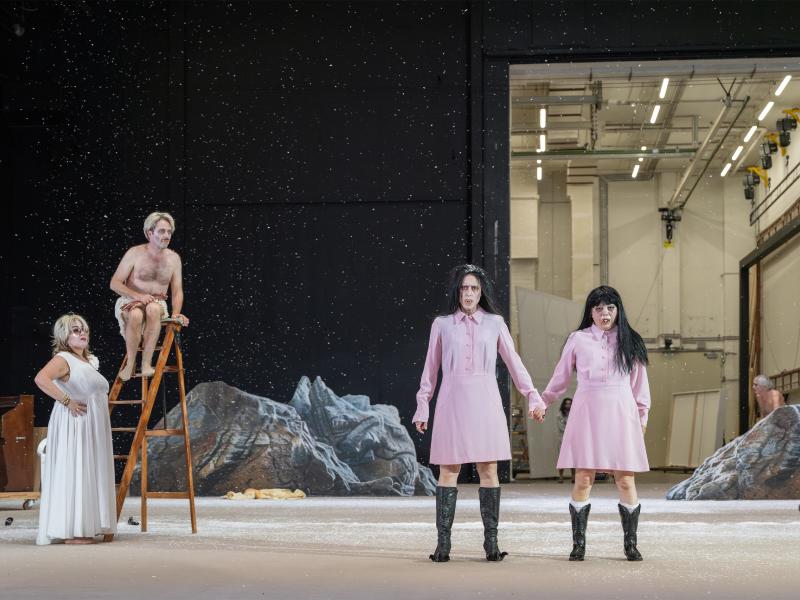 Sasha Rau und Sachiko Hara stehen Hand in Hand nebeneinander, sie tragen beide das gleiche rosa Kleid. Links von ihnen Bettina Stucky und Samuel Weiss, der auf einer Leiter sitzt und einen blutigen Arm in der Hand hält. Im Hintergrund ein Klavier, Felsen und ein offenes Portal zur Hinterbühne. 