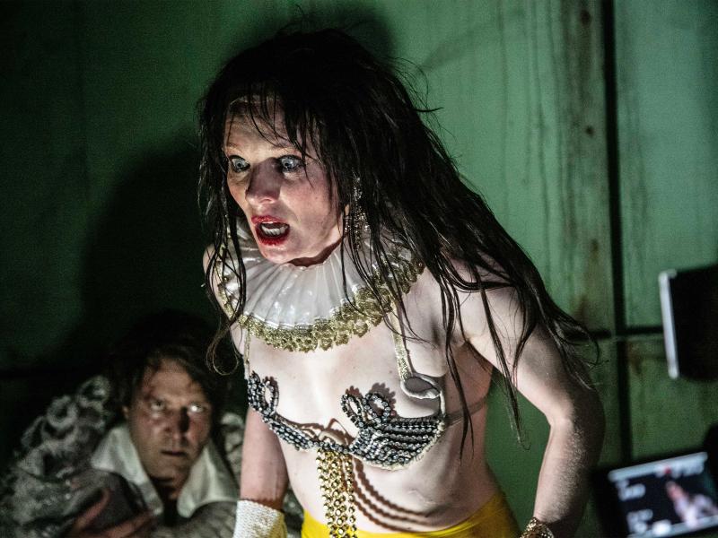 Lilith Stangenberg mit einer Perücke aus langen braunen Haaren, die verwuschelt sind. Sie trägt einen mit Glitzer besetzten knappen BH mit Verschnörkelungen und eine weiße Halskrause mit goldener Borte. Ihre Augen sind weit aufgerissen und ihr Mund geöffnet, sie trägt kräftigen roten Lippenstift.