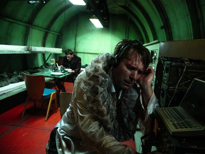 Ein geschlossener röhrenförmiger Raum mit Leuchtstoffröhren an der Decke. Im Vordergrund ein Mann mit silberner Jacke und Halskrause, er trägt Kopfhörer und sitzt vor einem schwarzen Monitor mit Tastatur, eine Hand am linken Ohr. Der zweite Mann sitzt im Hintergrund an einem grünen Klapptisch vor einem leeren Teller, vor ihm ein Topf.