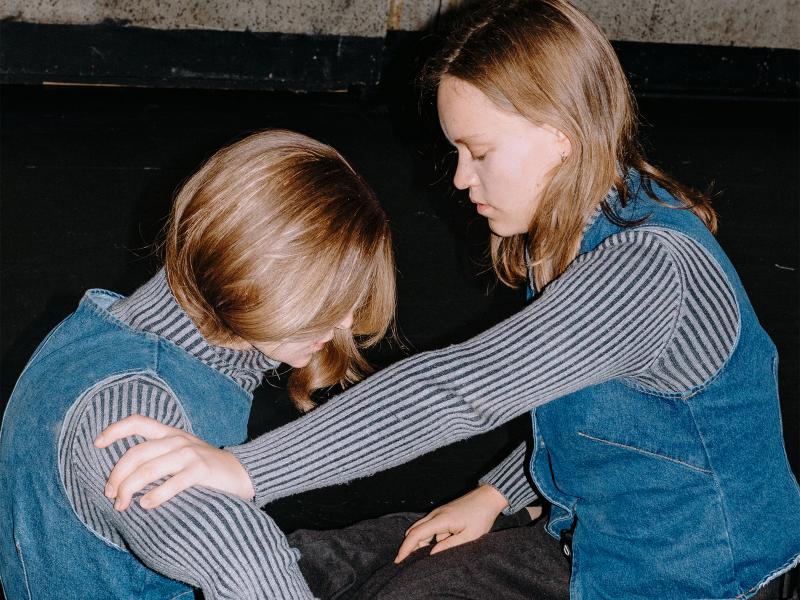 Lili Süper kniet auf dem Boden. Mit ihrem rechten Arm hält sie eine Puppe, die genau so aussieht wie sie (gleiche Größe, gleiche Kleidung, gleiche Haare). 