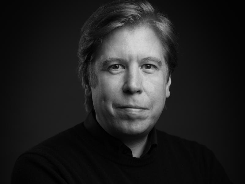 Schwarz-Weiß-Foto. Benjamin Sprick steht vor einem neutralen Hintergrund. Er trägt ein dunkles Hemd und schaut in die Kamera. Er schmunzelt leicht.