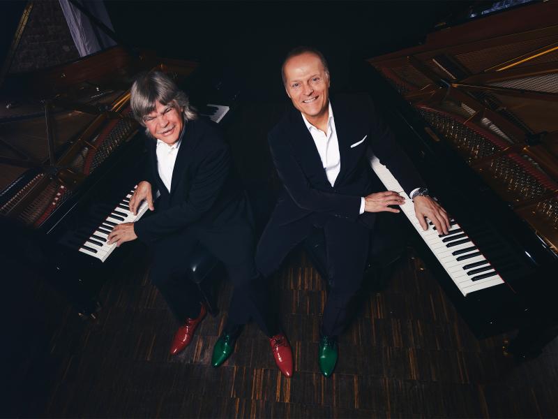 Die Pianisten Axel Zwingenberger und Joja Wendt sitzen jeweils an einem Flügel. Die Kamera ist in der Draufsicht leicht von oben platziert. Beide tragen ein weißes Hemd und einen dunklen Anzug. Sie lächeln in die Kamera.