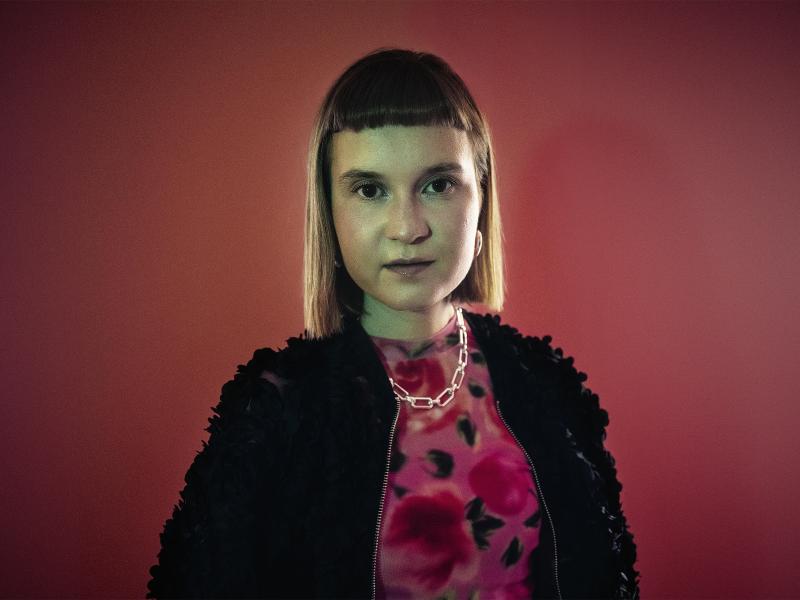 Caroline Wahl steht vor einem roten Hintergrund. Sie schaut in die Kamera. Sie trägt einen kinnlangen Bob und einen kurzen Pony. Sie hat ein pink-gemustertes Oberteil an, eine schwarze Jacke und eine silberne Kette.