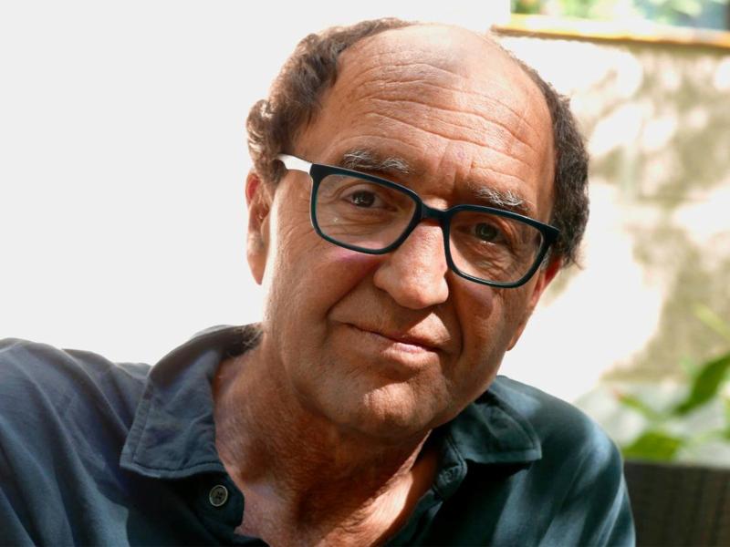 Doğan Akhanlı sitzt vor einer Hauswand. Er trägt ein blaues Hemd, kurze braune Haare und eine schwarze Brille. Er schaut in die Kamera.