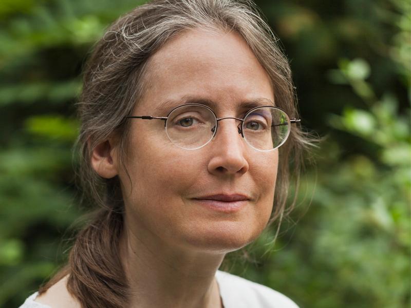 Porträt. Isabel Fargo Cole steht draußen in der Natur, hinter ihr sind grüne Blätter zu sehen. Sie steht etwas schräg zur Kamera und schaut hinein. Sie trägt zusammengebundenes, schulterlanges Haar und eine filigrane Brille.