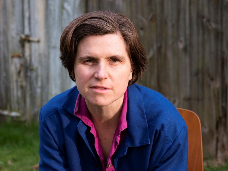 Judith Schalansky sitzt vor einer hölzernen Wand. Sie trägt ein pinkes Hemdn und einen blauen Blazer. Sie hat kinnlanges braunes Haar und schaut in die Kamera.