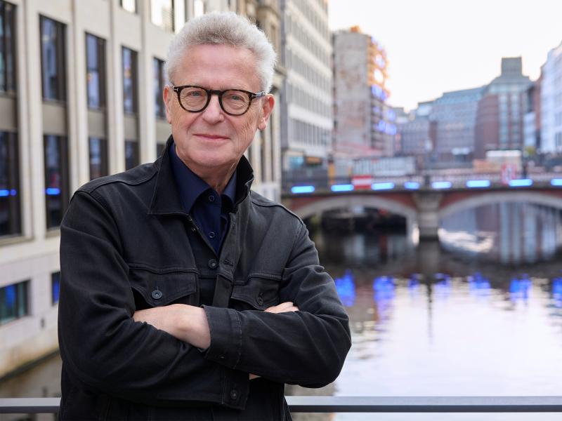 Michael Batz trägt eine schwarze Jacke, ein dunkelblaues Hemd und eine dunkle Brille. Im Hintergrund ist ein Hamburger Fleet und Gebäude der Stadt in der Unschärfe zu sehen.