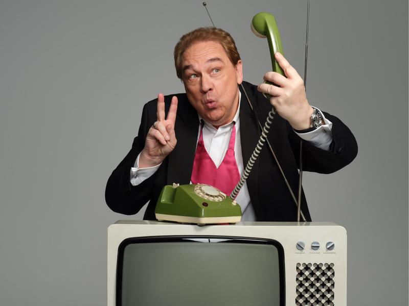 Oliver Kalkhofe steht vor einem grauen Hintergrund. Er trägt ein weißes Hemd, eine pinke Weste und ein schwarzes Sakko. Vor ihm steht ein alter Fernseher. Er hebt den Hörer eines alten Drehscheibentelefons mit Kabel in die Höhe, schaut ihn an und macht ein "Peace"-Zeichen mit Zeige- und Mittelfinger. Er macht eine Grimasse zum Telefonhörer, als würde er ein Foto von sich selber machen wollen.