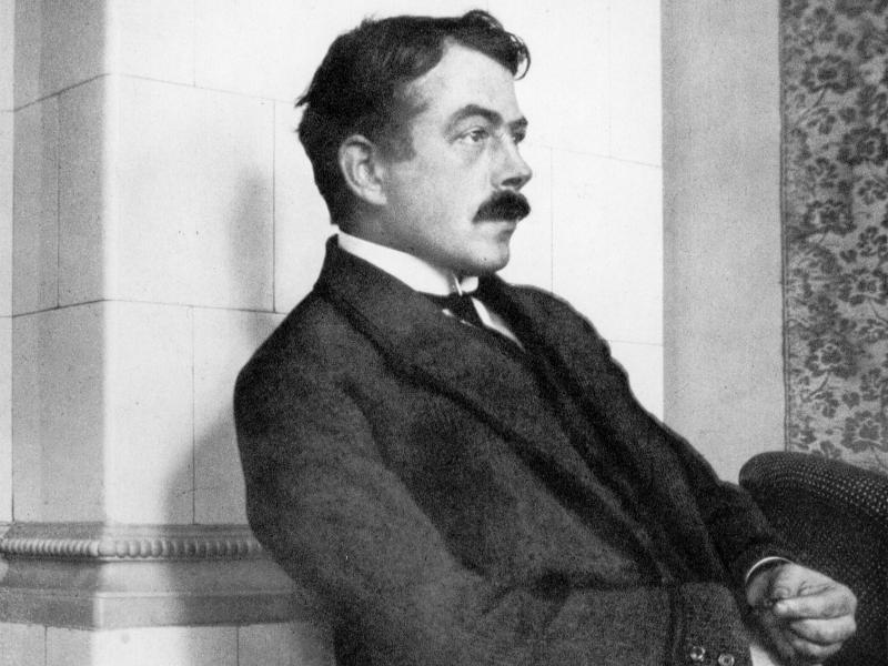 Schwarz-Weiß_Foto. Robert Walser sitzt im Anzug vor einer weißen Wand.