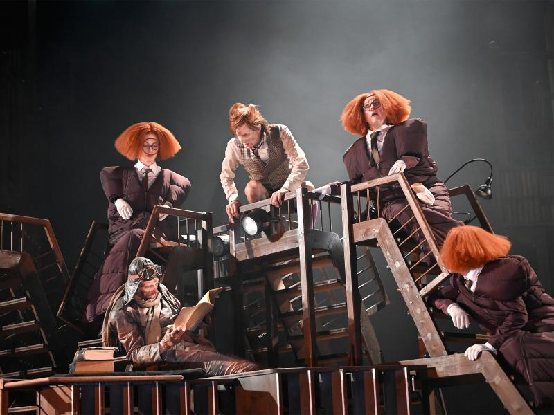 Auf einer Ansammlung von Treppen hockt Jan-Peter Kampwirth und blickt nach unten zu Jan Thümer mit Pilotenmütze, der in einem Manuskript liest. Ebenfalls auf den Treppen sitzen drei Personen mit aufgeplusterten orangenen Perücken. Sie blicken in alle Richtungen.