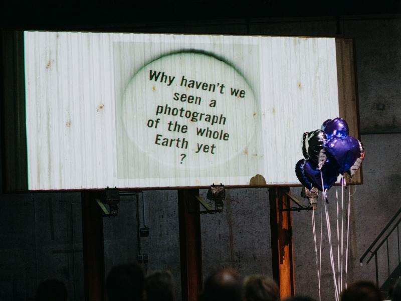 Auf der Bühne des MalerSaals steht eine Leinwand, auf die ein Bild von einem Kreis projiziert wird. In dem Kreis steht: „Why haven’t we seen a photograph of the whole Earth yet?“. Rechts daneben sind silberne und blaue herzförmige Heliumluftballons. 