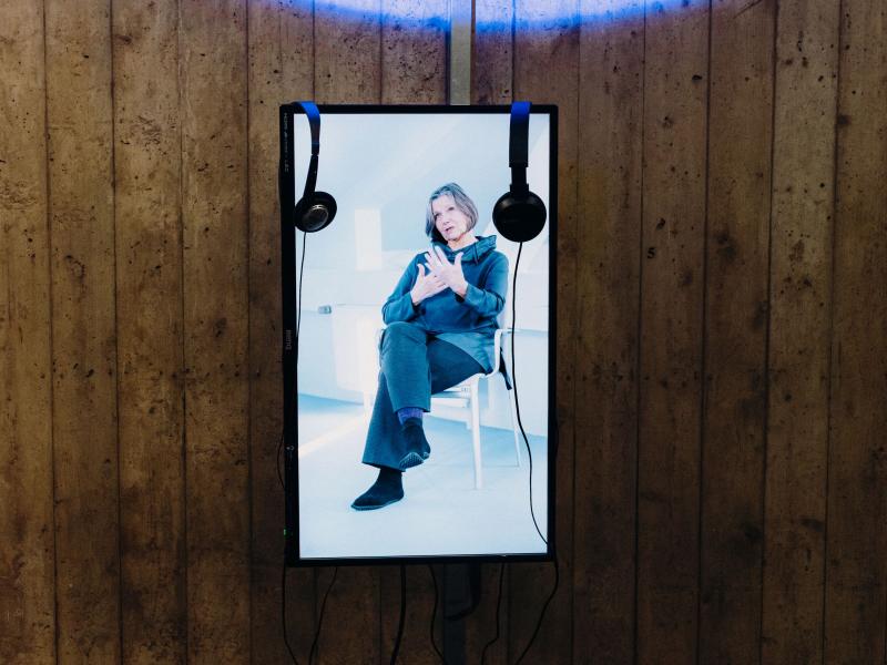 Es ist ein Bildschirm zu sehen, der eine Frau zeigt. Sie sitzt auf einem Stuhl und gestikuliert mit ihren Händen. An dem Bildschirm hängen zwei Kopfhörer.