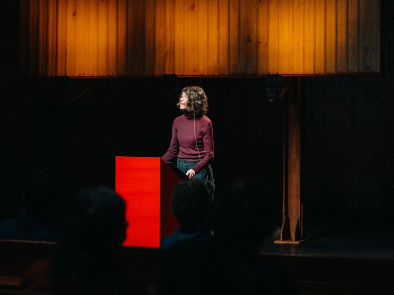 Louise Brown steht an einem roten Podium und spricht. Der Raum ist abgedunkelt nur sie ist beleuchtet. 