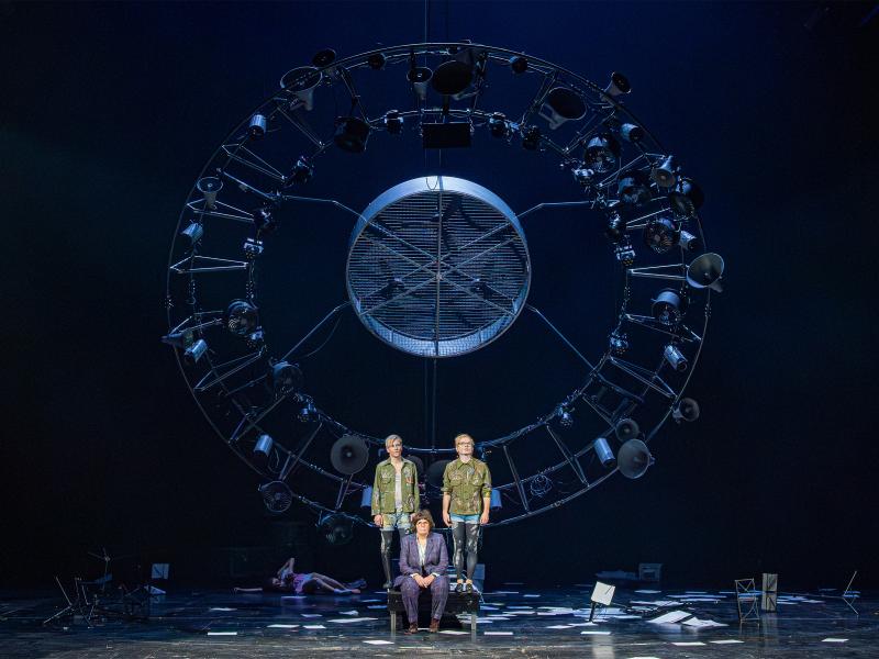 Eine senkrecht hängende große runde Stahlkonstruktion aus vielen unterschiedlich großen alten trichterförmigen Lautsprechern, Scheinwerfern und Ventilatoren. Davor stehen Kristof Van Boven und Nils Kahnwald auf einem Podest. Sie tragen schwarze Leggings mit weißen Mustern, kurze Jeanshosen darüber und dunkelgrüne Jacken mit Symbolen und Bildern, wie einem Spinnennetz, Sternen, Feuer, Herzen, Skeletten. Zu ihren Füßen sitzt Julia Wieninger in einem lila-grün karierten Anzug. Auf dem Boden verteilt