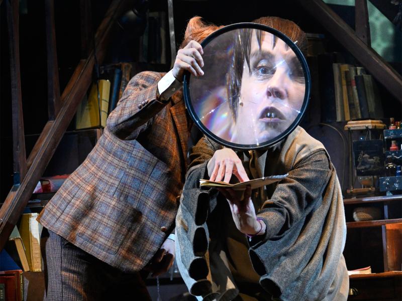 Jan-Peter Kampwirth und Ute Hannig stehen dicht nebeneinander und blicken durch ein großes Vergrößerungsglas, sodass vor allem Ute Hannigs Gesicht stark vergrößert ist. In den Händen hält sie ein Manuskript.