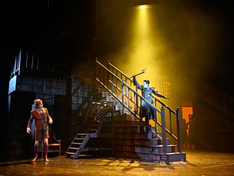 Ute Hannig steht auf einer Treppe eines großen Bücherregals und streckt eine lange Axt in die Höhe. Jan-Peter Kampwirth guckt sie von unten an. 