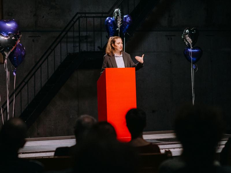 Prof. Anna-Verena Nosthoff steht an einem roten Podium und gestikuliert mit ihrer Hand. Um sie herum sind silberne und blaue herzförmige Heliumluftballons.