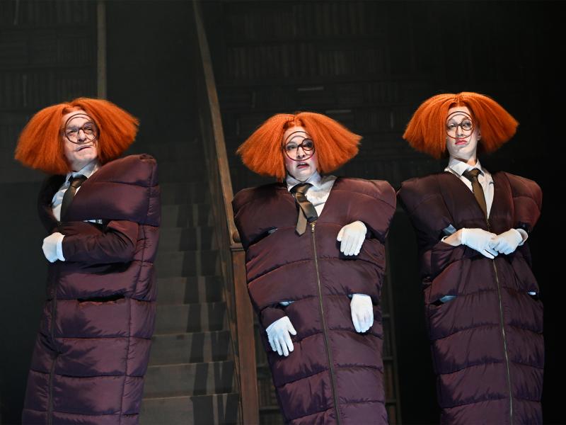 Drei Personen in Daunenjacken-Kleidern und mit orangen aufgeplusterten Perücken stehen nebeneinander vor einem großen Bücherregal. Die mittlere Person hat drei Hände. Sie blicken mit verschiedenen Gesichtsausdrücken in die Ferne.