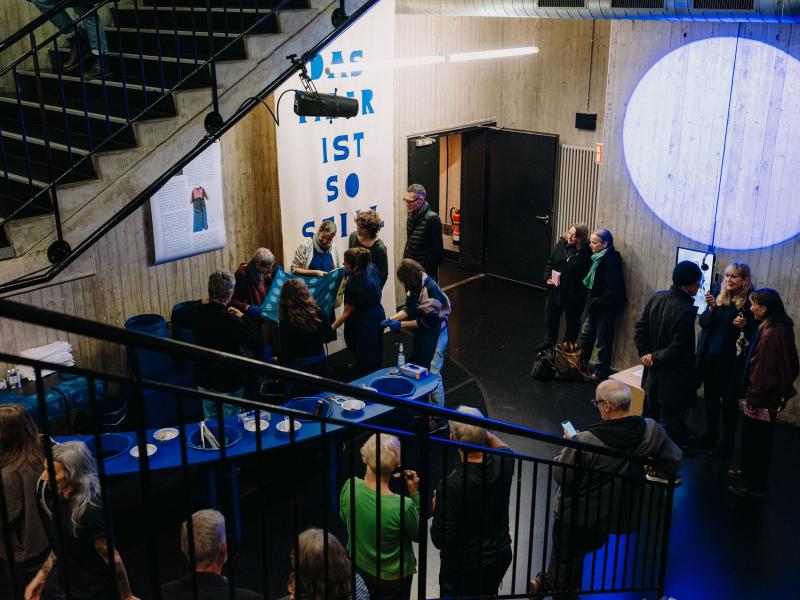 Viele Menschen stehen verteilt im MalerSaalFoyer, einige halten blau gefärbte Tücher in den Händen. In der Mitte des Raumes ist ein blauer langer Tisch, auf dem sich Werkzeuge und Bastelutensilien befinden. Es sind Eimer in den Tisch eingelassen. 