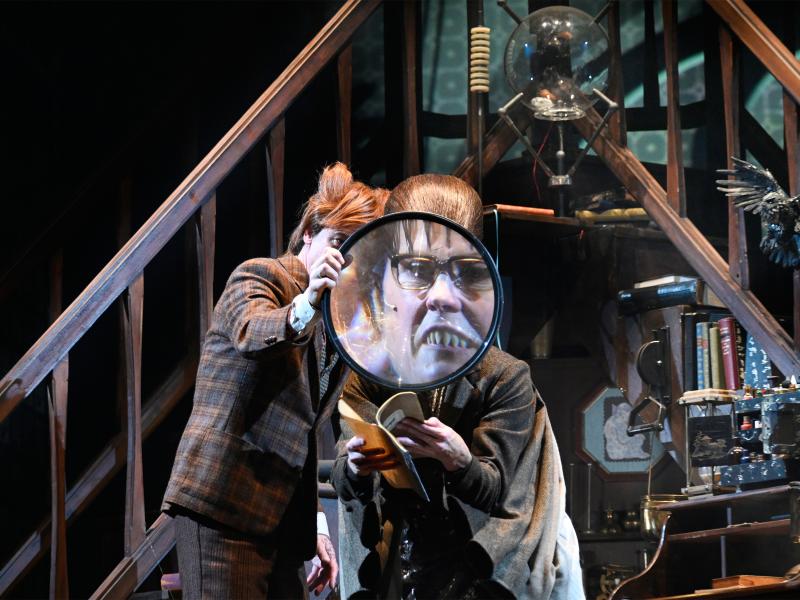 Jan-Peter Kampwirth und Ute Hannig stehen vor zwei gegenüber zulaufenden Treppen in einem eingerichteten Büro, mit vielen wissenschaftlichen Instrumenten. Jan-Peter hält eine überdimensionale Lupe vor Utes Gesicht, wodurch man es vergrößert sieht.