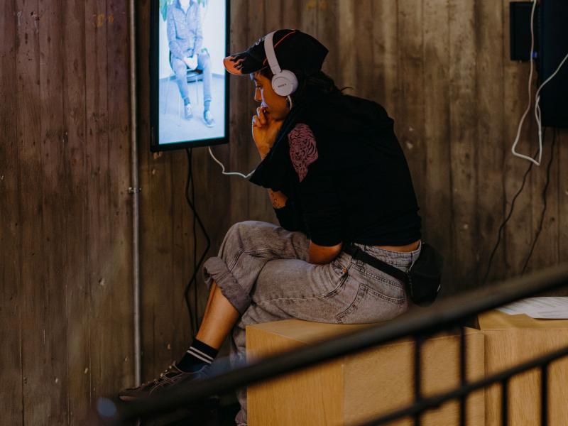 Eine Person sitzt in der Ecke des MalerSaalFoyers auf einem Hocker. Sie trägt einen Kopfhörer, mit dem sie etwas aus der Ausstellung hört. Die Kopfhörer sind an einen kleinen Monitor befestigt, auf der eine Person halb zu erkennen ist.