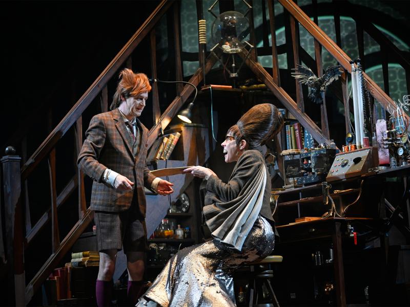 Jan-Peter Kampwirth steht vor zwei gegenüber zulaufenden Treppen in einem eingerichteten Büro, mit vielen wissenschaftlichen Instrumenten. Neben ihm sitzt Ute Hannig auf einem Stuhl und hält ihm eine Mappe hin. 