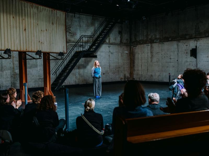 Lili Süper steht auf der leeren Bühne des MalerSaals mit den Armen hinter den Rücken verschlossen und lächelt. Das Publikum ist von hinten zu sehen, es klatscht.