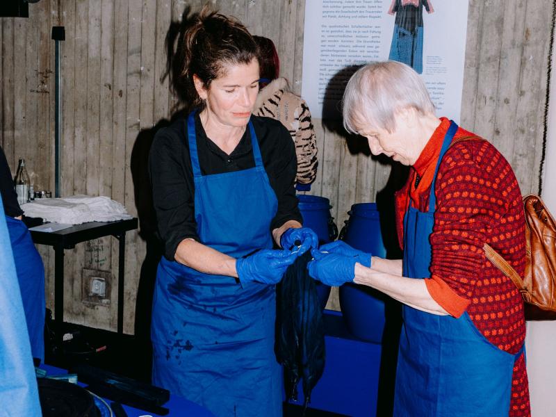 Zwei Frauen in blauen Schürzen und mit blauen Handschuhen halten im MalerSaal Foyer blau gefärbte Tücher in den Händen. 