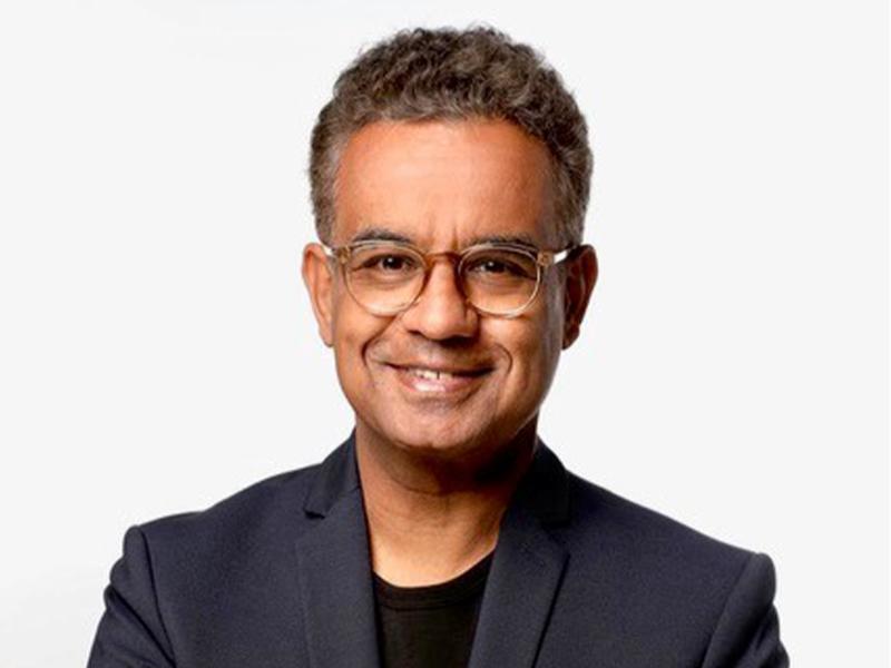 Michail Paweletz steht vor einer weißen Wand. Er trägt ein schwarzes T-Shirt, darüber ein dunkles Sakko und eine helle, runde Brille. Er lächelt und schaut in die Kamera.