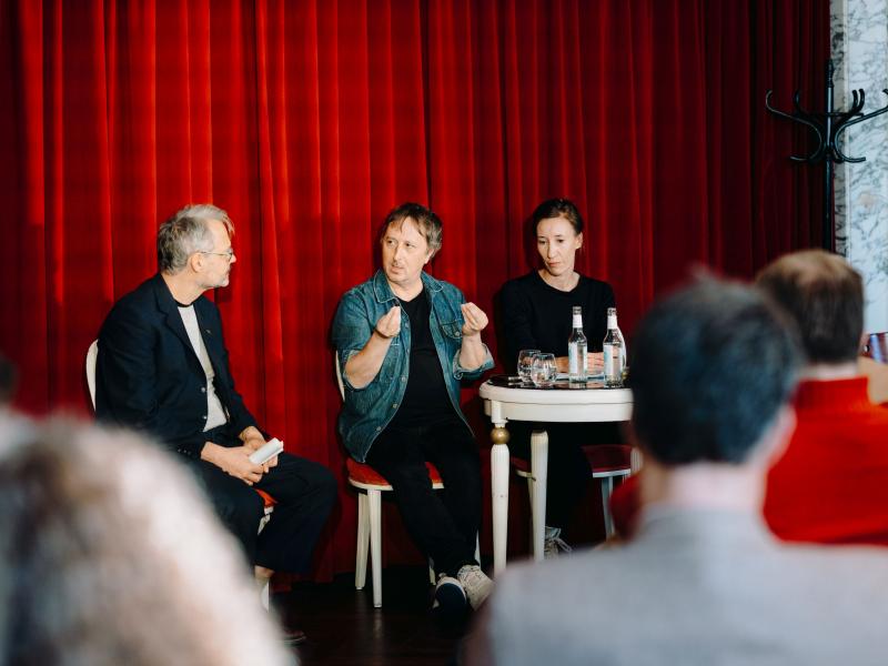 Christian Tschirner, Phillippe Quesne und eine Frau sitzen gemeinsam an einem Tisch im MarmorSaal. Phillippe Quesne gestikuliert mit seinen Händen, die anderen schauen ihn an. Das Publikum ist ebenfalls von hinten zu sehen.