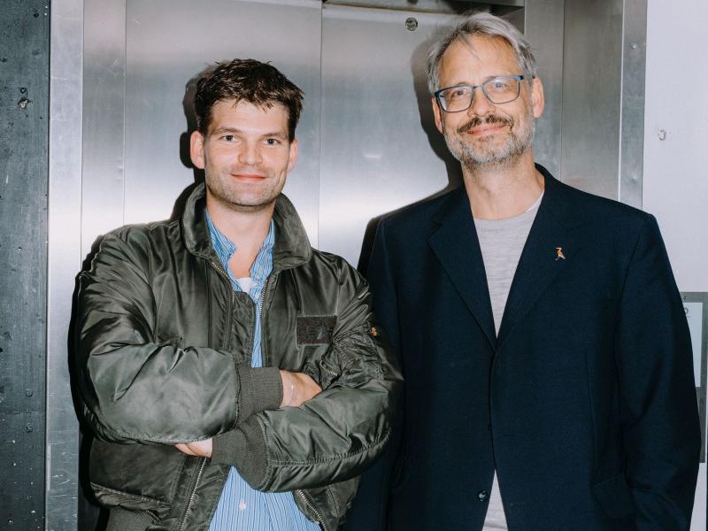 Martin Györffy und Christian Tschirner stehen nebeneinander vor einem Fahrstuhl und lächeln. 