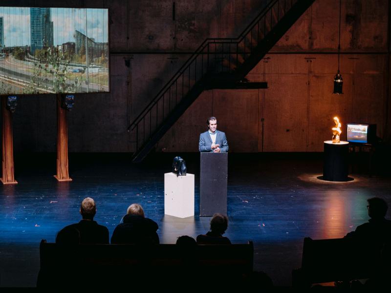 Wilson Tanner Smith steht auf der Bühne des MalerSaals an einem Podium. Im Hintergrund ist eine Leinwand zu sehen, auf der ein Foto von einer Autobahn und Hochhäusern abgebildet ist. Die im Publikum sitzenden Menschen sind von hinten zu sehen.